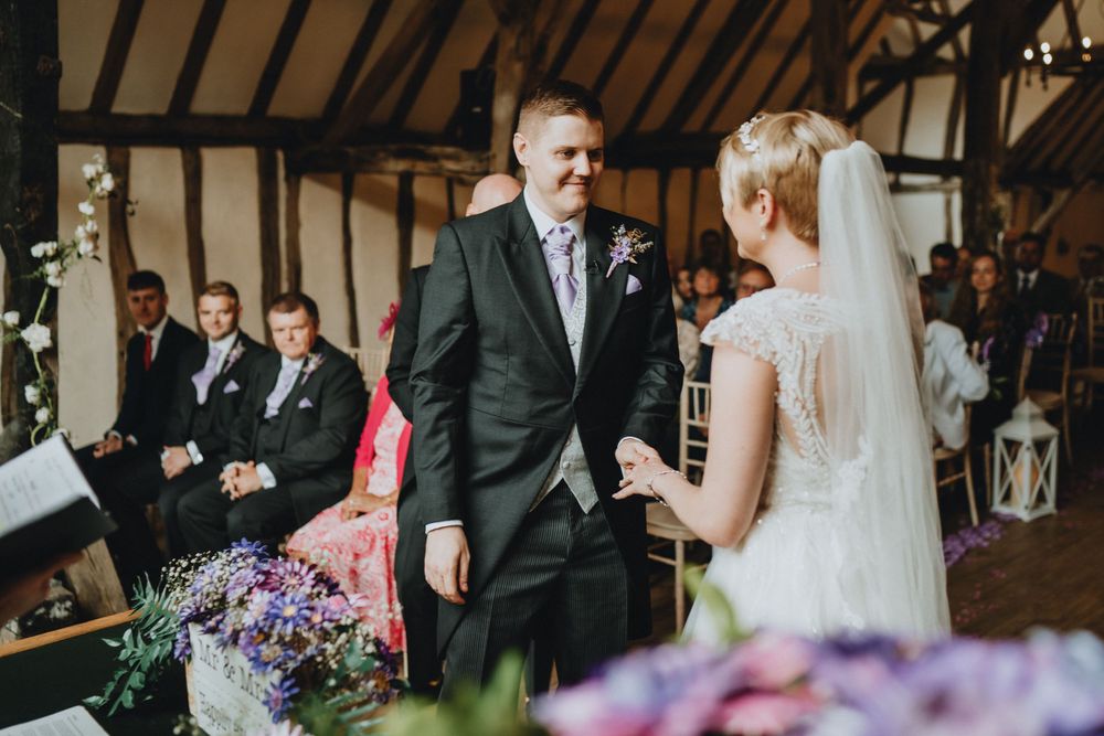 Two people exchange vows during a rustic barn wedding ceremony with purple floral decorations in the foreground.