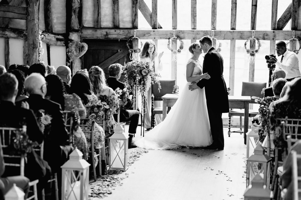 A black and white photo of a wedding ceremony in a rustic barn lined with lantern-lit aisle decor.