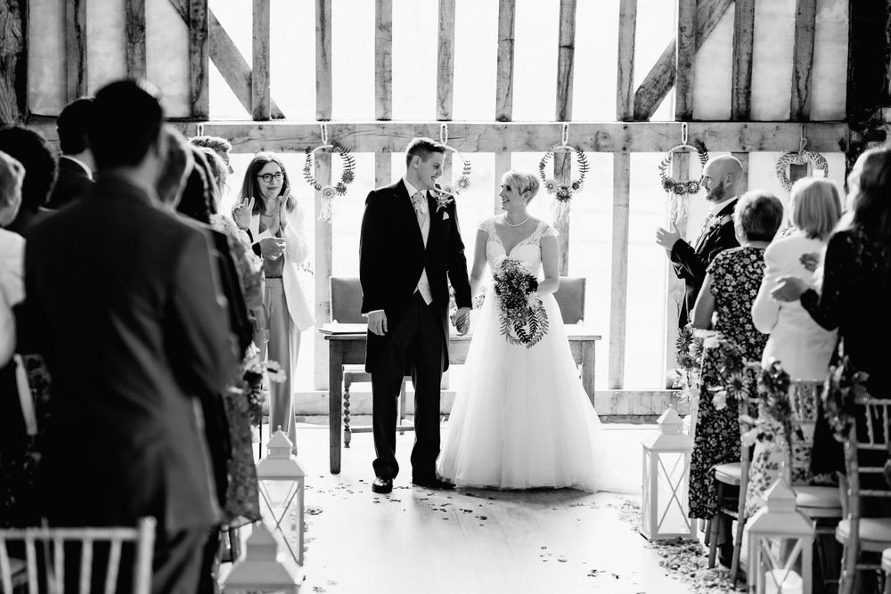 A black and white photo of a wedding ceremony inside a timber-framed barn with guests seated on either side.