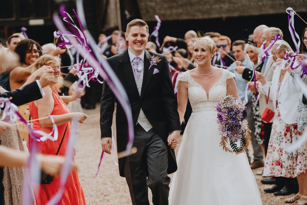 Wedding guests wave colorful ribbon wands as newlyweds walk through crowd during outdoor celebration.