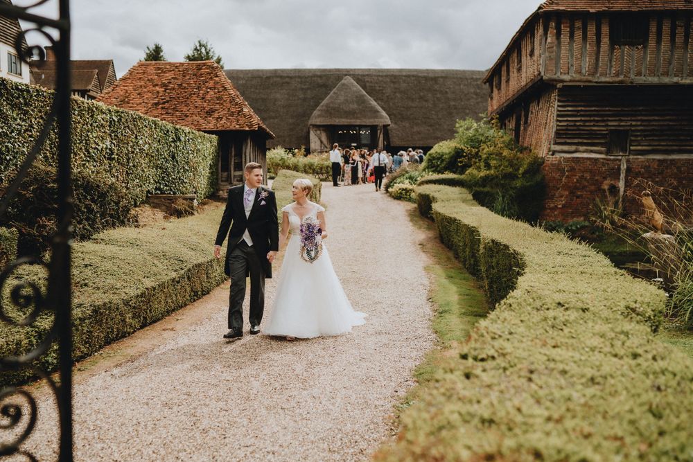 Newlyweds walking down rustic garden path between hedge rows at countryside wedding venue.