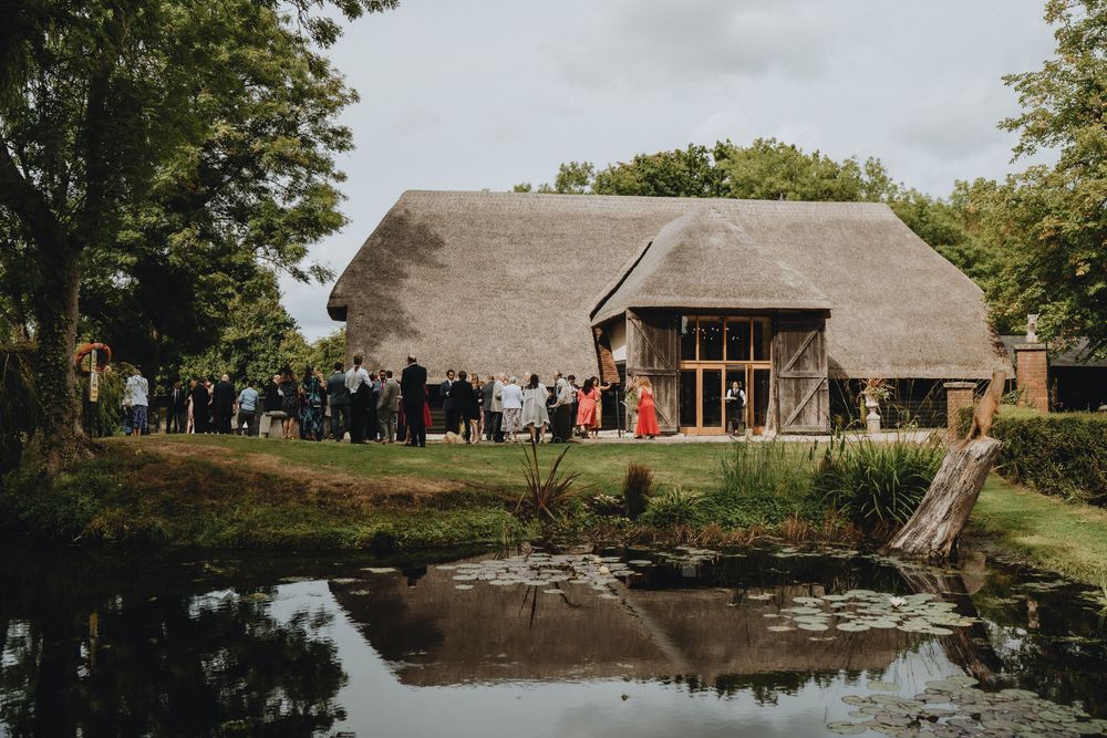 A rustic thatched-roof barn venue with guests gathered outside of Colville Hall beside a reflective pond on a rural property.