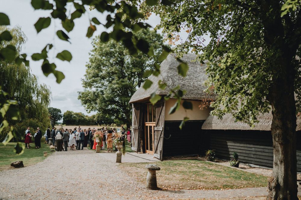 Wedding guests gather outside a rustic barn venue with trees and a gravel path on a sunny afternoon.