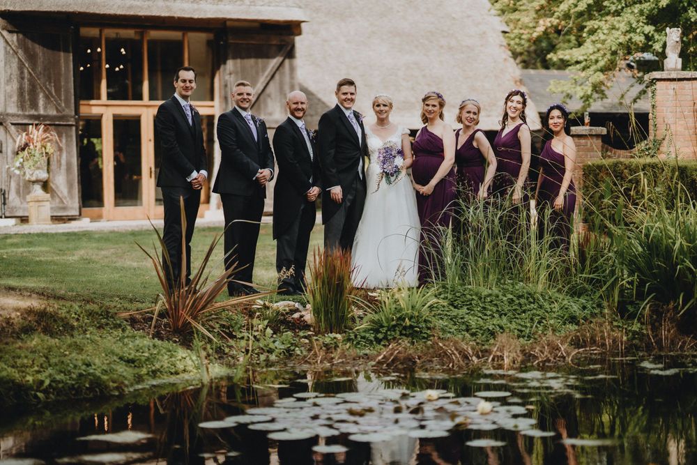 Wedding party stands in elegant formation by a reflective pond at a rustic venue with wooden barn structure.