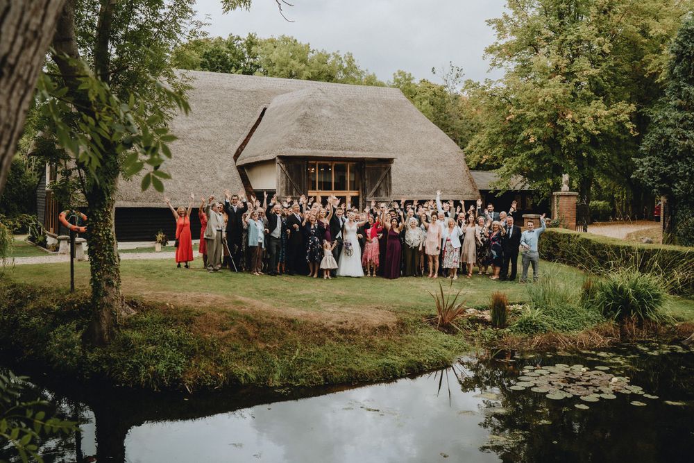 Large group of wedding guests posed in front of a rustic barn next to a pond on a beautiful summer day.
