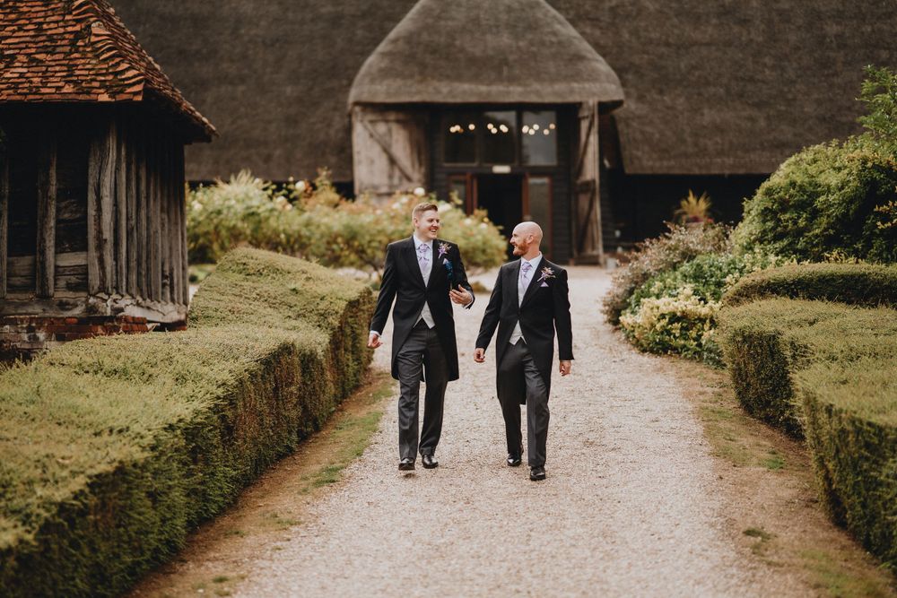 Two people in formal attire walking down a garden path towards a rustic barn entrance.