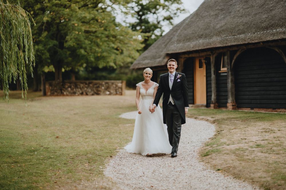 A newlywed couple walks together on a gravel path near a rustic barn building with a willow tree in the background.