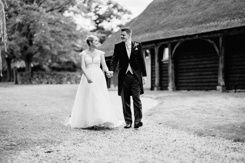 Black and white wedding photograph showing couple walking together outside a rustic barn building with thatched roof.
