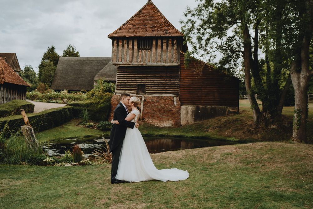 A couple embraces on a green lawn in front of a rustic brick barn with timber-framed tower on a cloudy day.