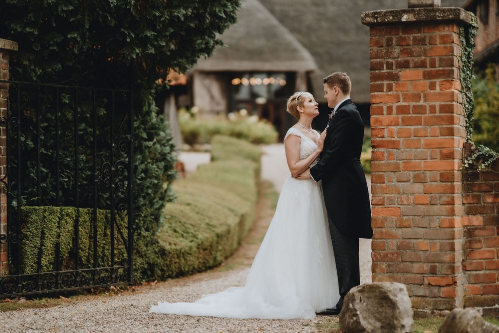 A romantic wedding moment on a rustic countryside path beside a red brick pillar at an English estate.