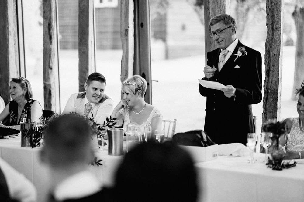 A black and white candid shot of wedding reception guests laughing while someone gives a speech at the event.