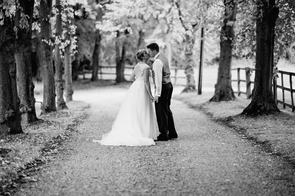 A romantic black and white photo of a couple embracing on a tree-lined path on their wedding day.