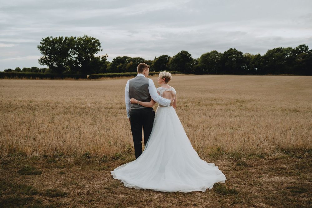 A couple in wedding attire stands together in a golden wheat field under an overcast sky.
