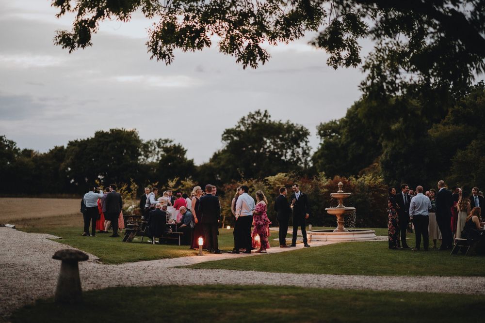 Wedding guests gather at an outdoor reception venue at dusk under towering trees.