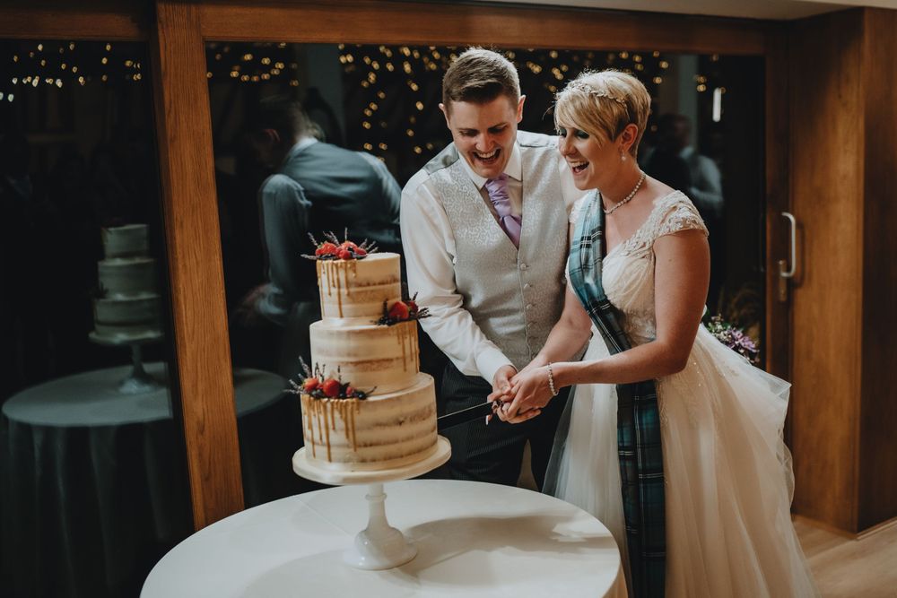 Couple cutting into their semi-naked wedding cake with dripping caramel and fresh flowers at rustic reception venue.