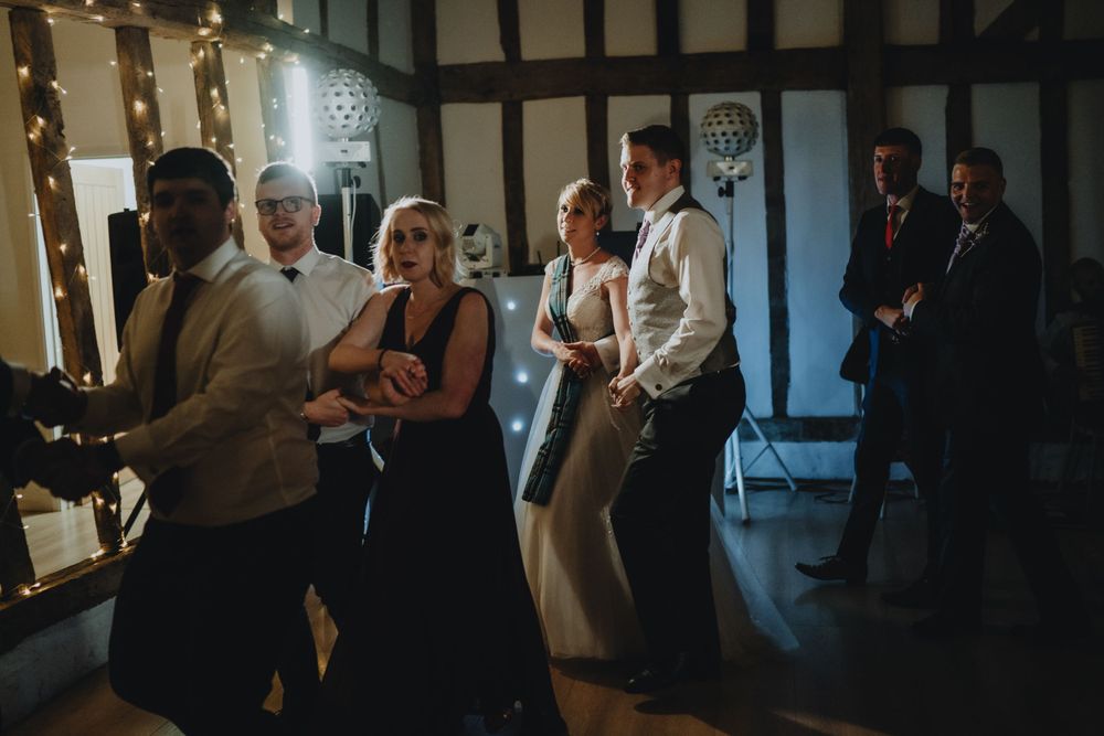 Wedding guests dancing and socializing at a reception in a rustic Tudor-style venue with exposed beams.