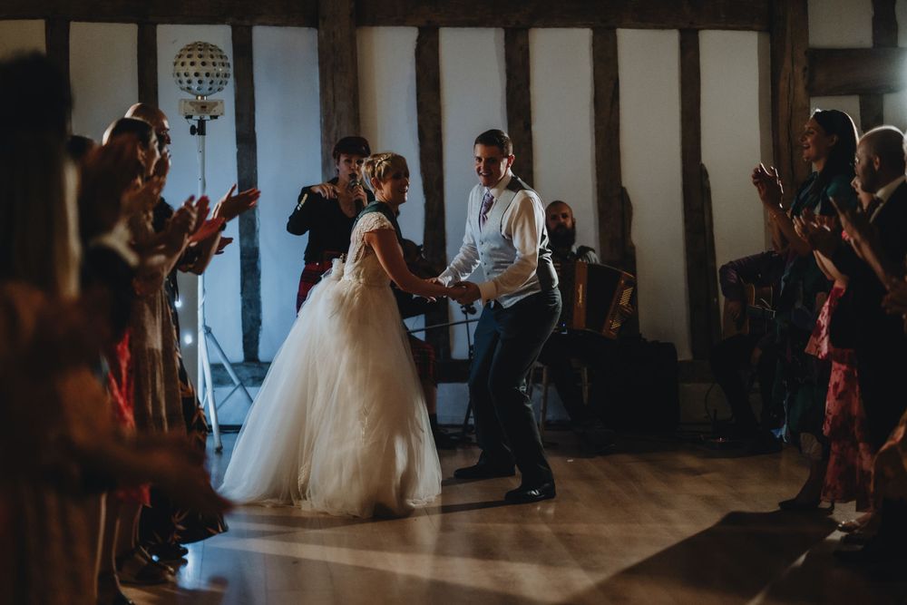 A couple shares their first dance at a wedding reception while guests applaud in a rustic venue with wooden beams.