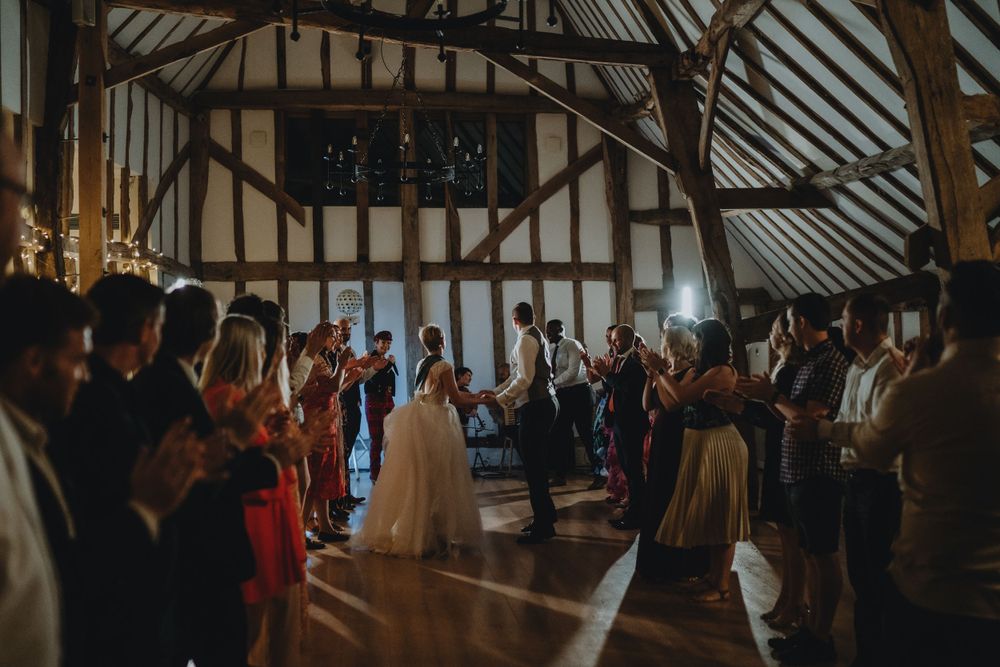Wedding guests celebrate and dance together inside a rustic timber-framed barn venue with dramatic lighting.