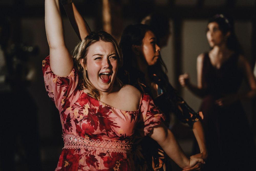 Dancing guest in red floral dress raises arm joyfully at dimly lit party celebration.