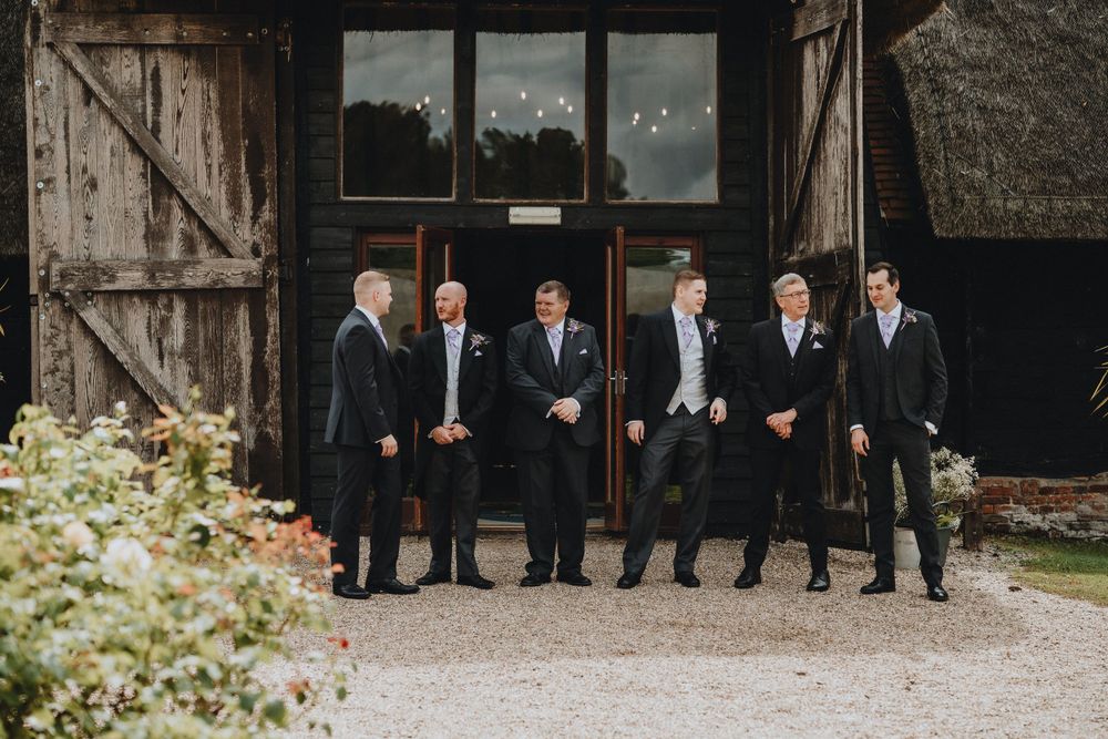 A group of groomsmen in dark suits pose together outside a rustic barn venue at Colville Hall.