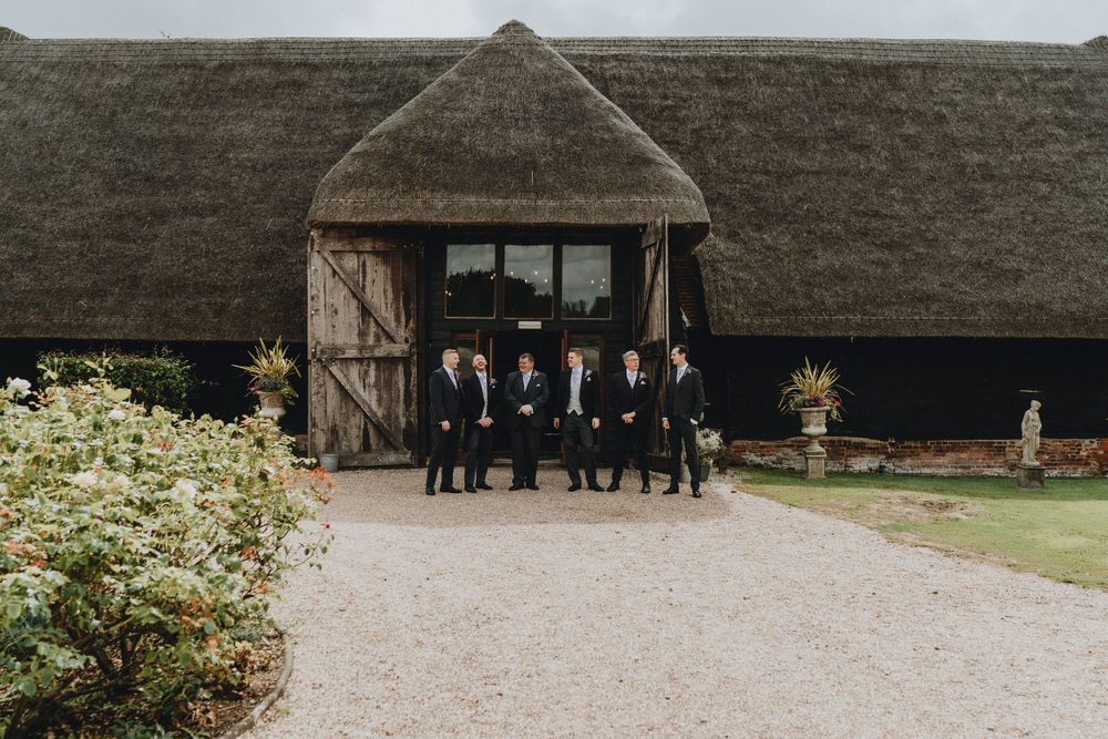 A group stands beneath a rustic thatched-roof entrance of a barn at Colville Hall on a gravel path.