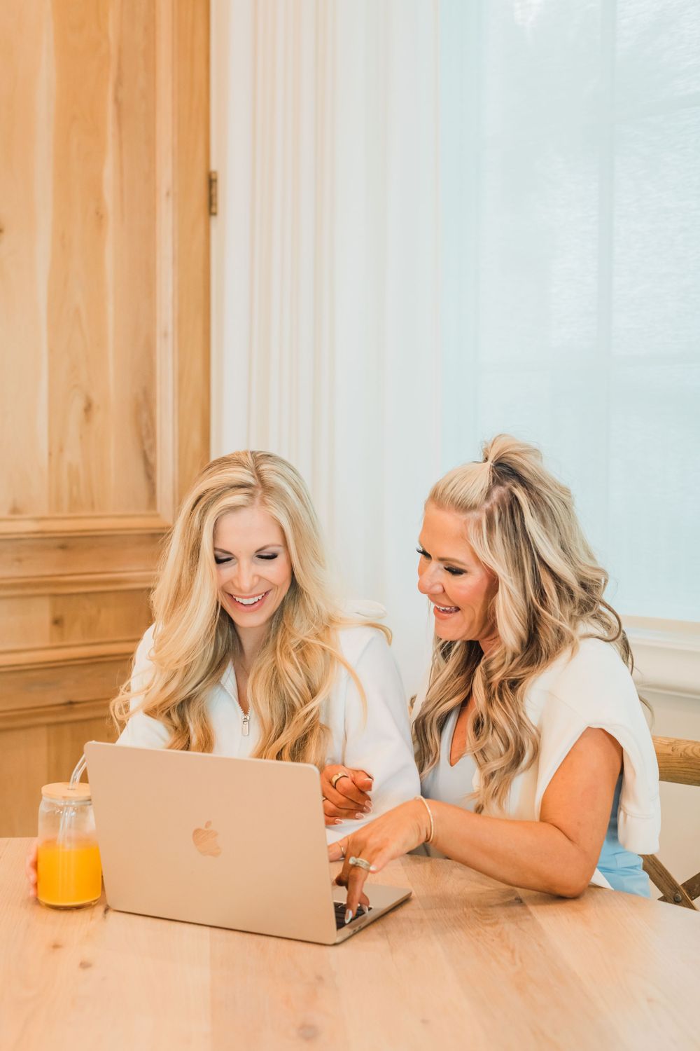 Two blonde women smiling while working together on a MacBook laptop at wooden table with orange juice nearby.