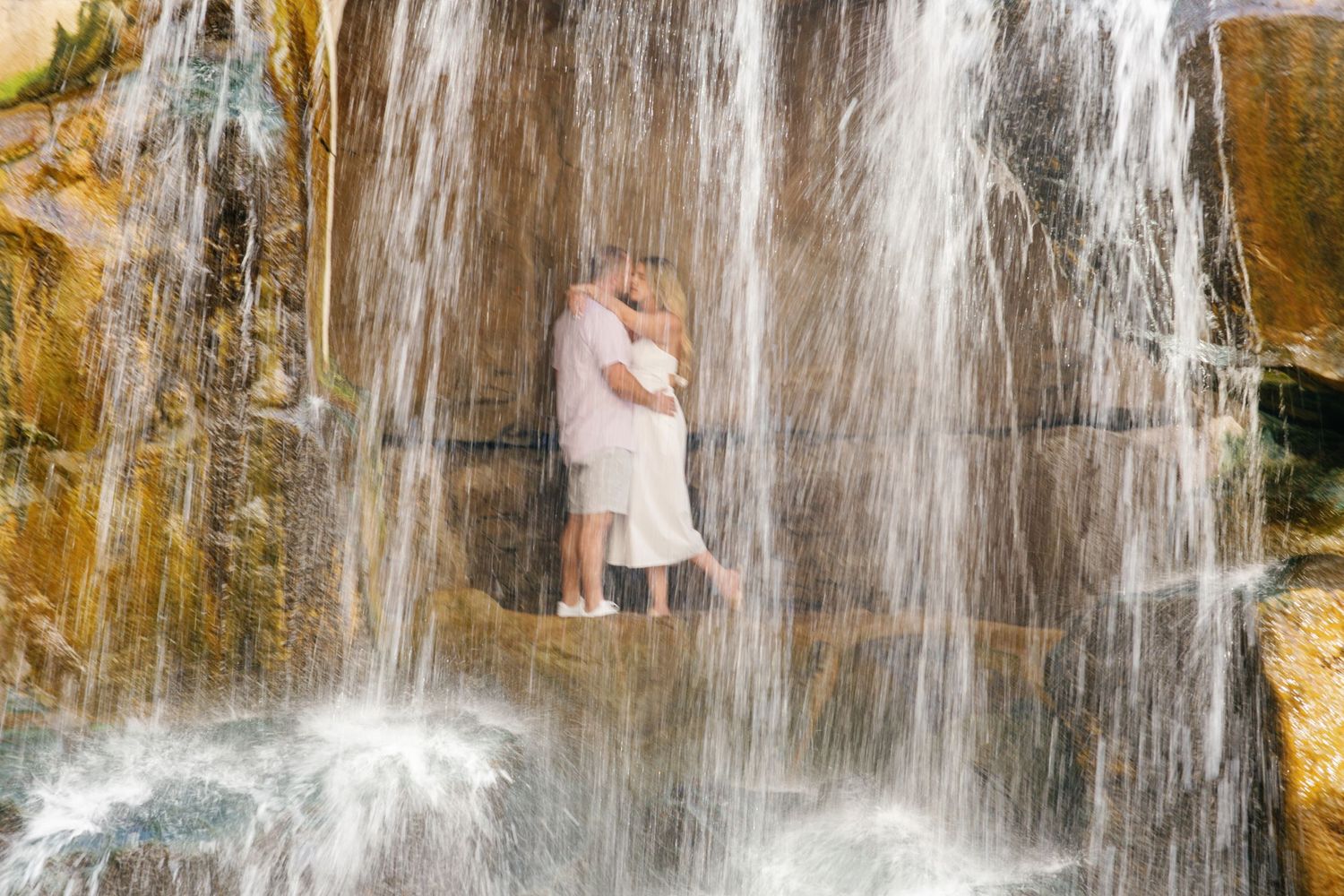 Two figures embrace under cascading waterfall with rocky walls and flowing water creating a romantic natural backdrop.