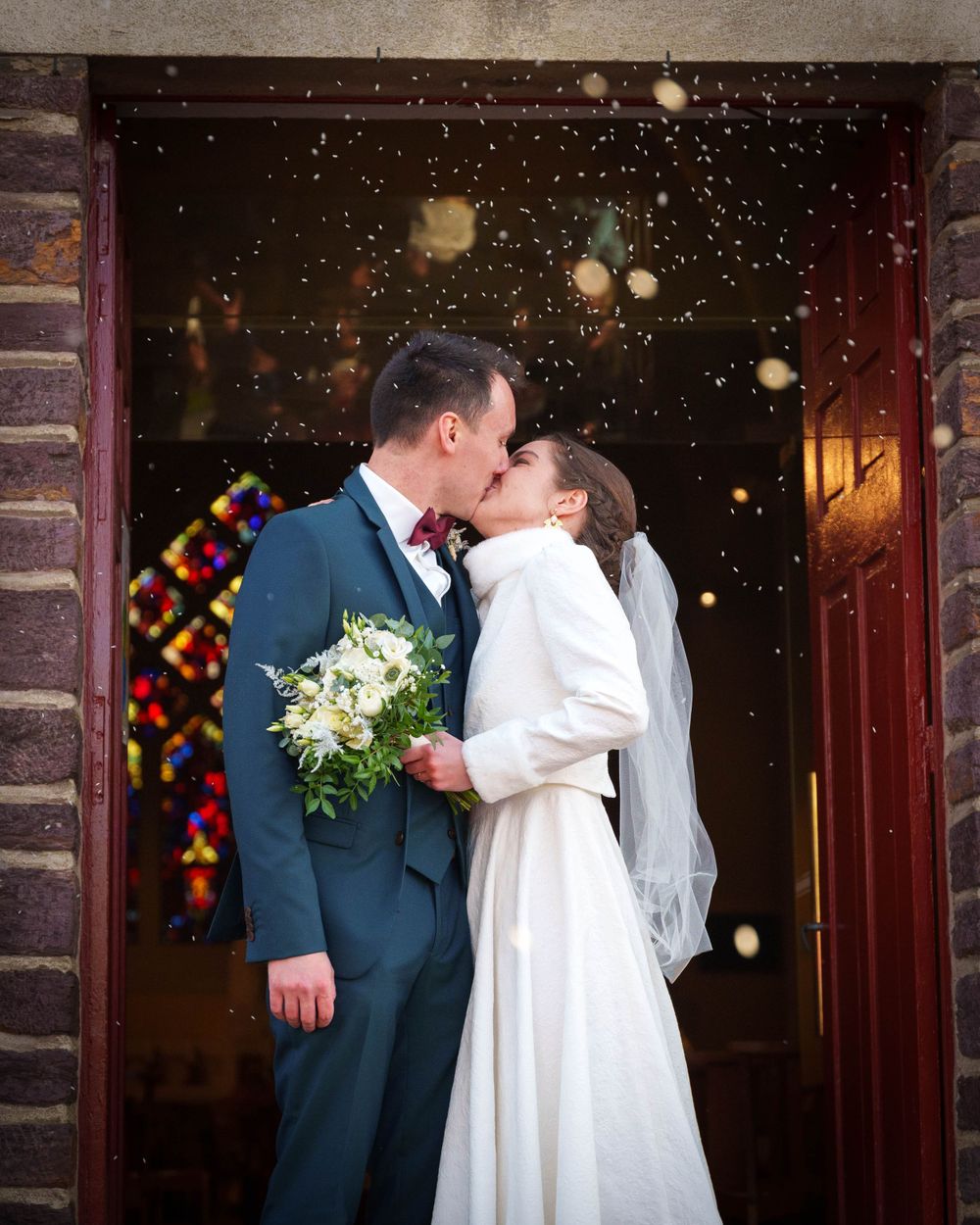 Deux jeunes mariés s'embrassent à la sortie de l'église après leur cérémonie de mariage. La mariée tient un bouquet dans la main.