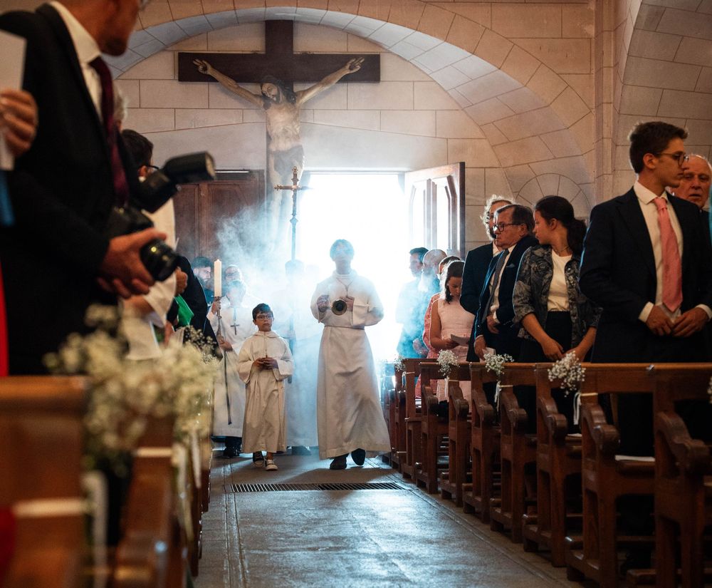 Wedding guests seated in wooden church pews with floral arrangements lining the aisle during a ceremony.