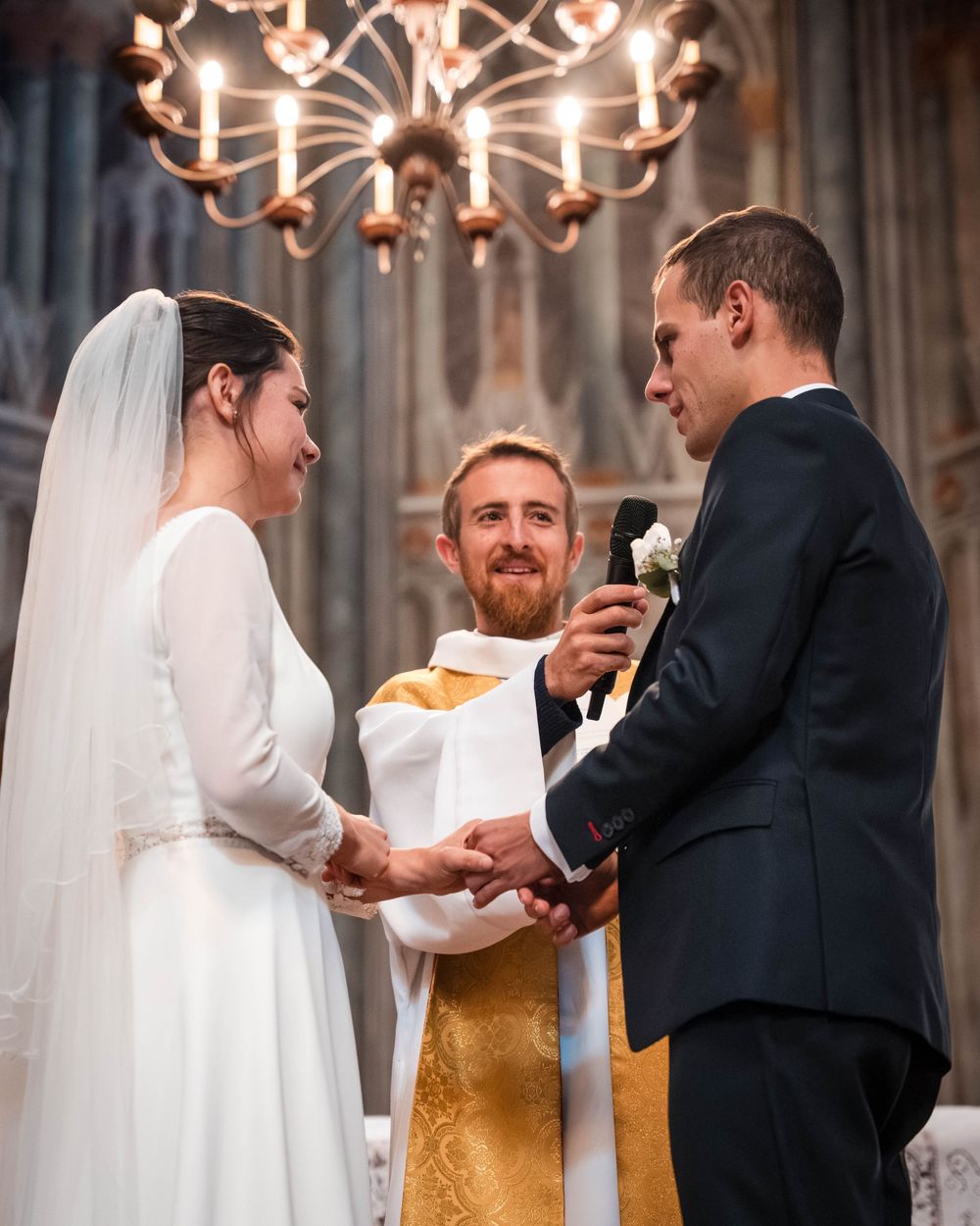 A couple exchanges rings during their wedding ceremony in a church under elegant chandeliers.
