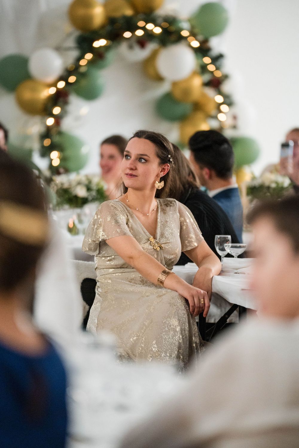 Une invitée vêtue d'une robe beige scintillante est assise à une réception élégante sous une arche décorée de ballons et de guirlandes