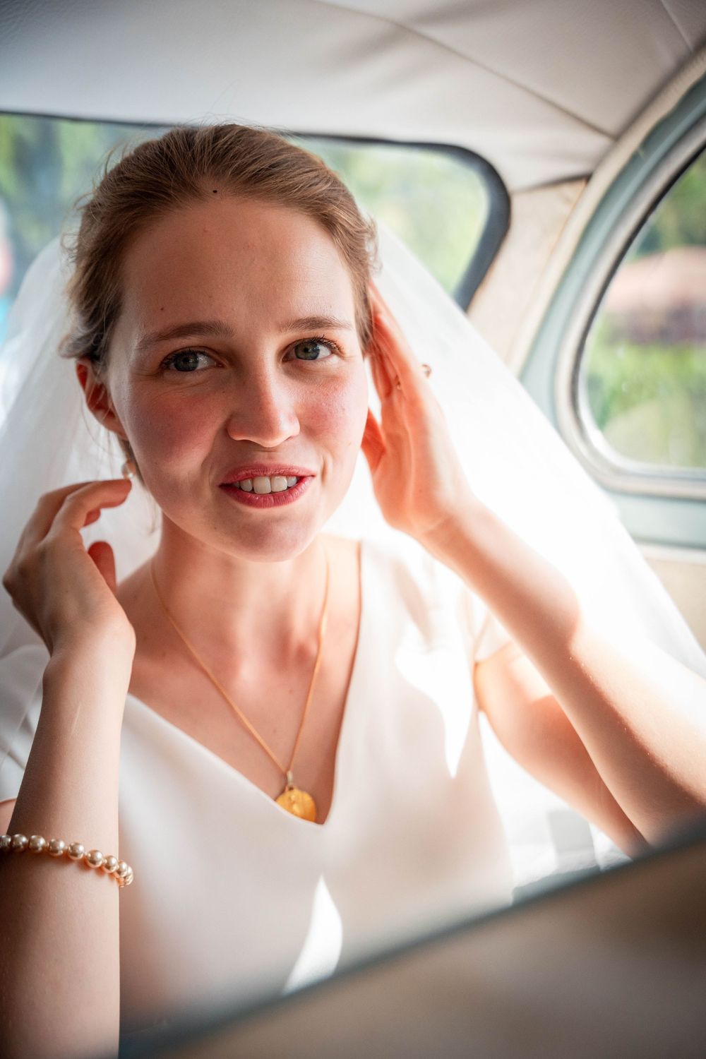 A smiling bride in a white dress and veil adjusts her hair while sitting in a vintage car on her wedding day.