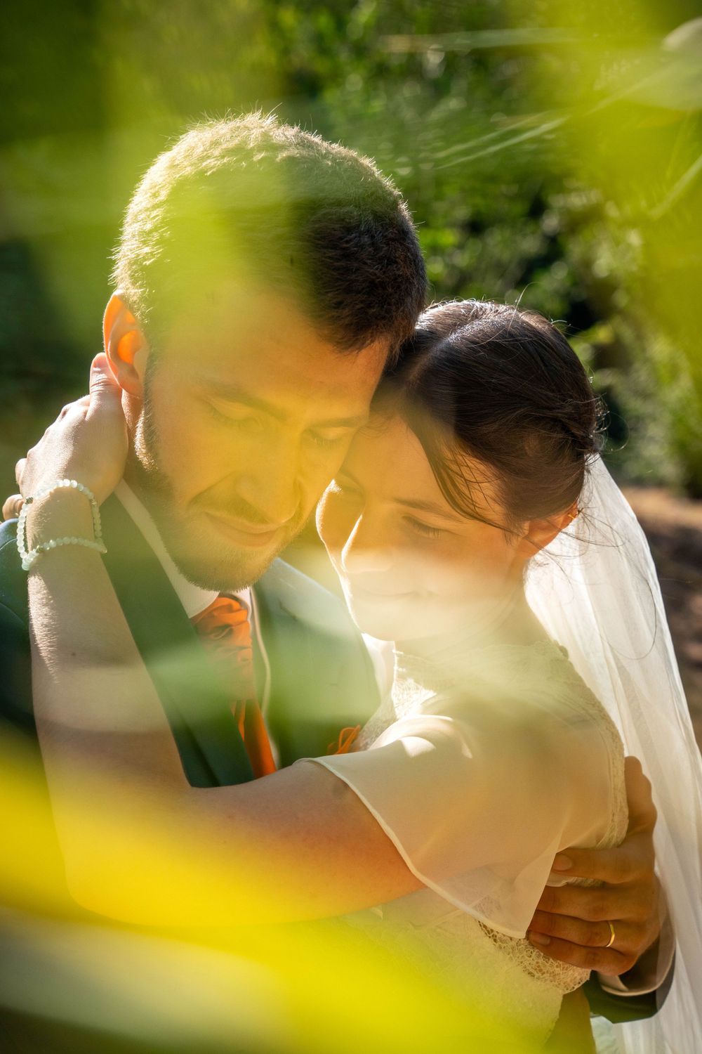 Une photo de mariage artistique sous un soleil doré avec un couple enlacé, encadré par un feuillage vert qui se superpose.