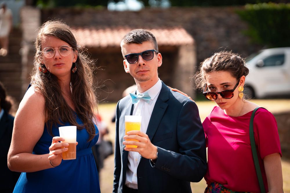 Three young adults holding drinks outdoors at a sunny event, dressed in formal attire including a suit and bow tie.