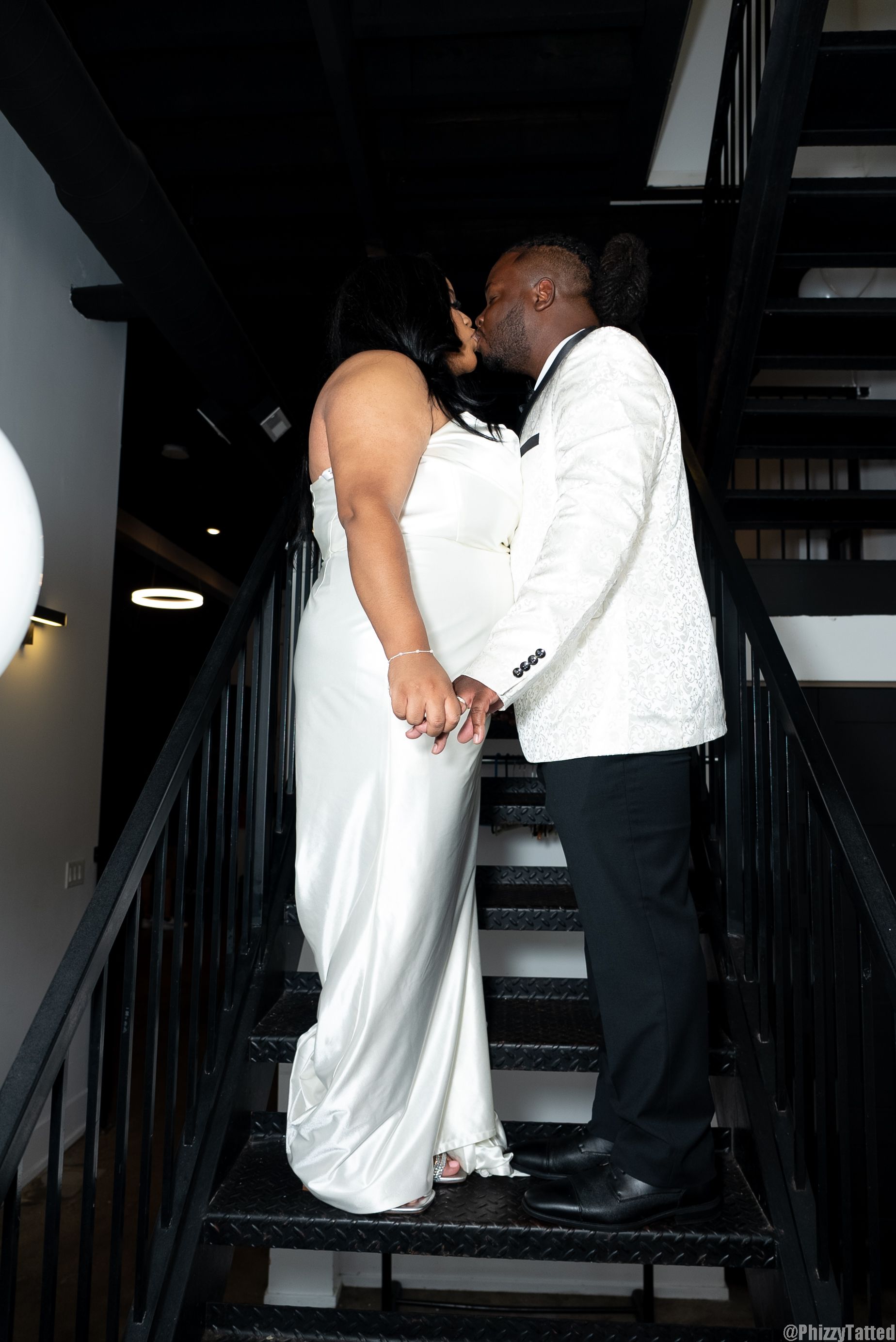 Couple sharing a romantic moment on black stairs with the person in white dress and person in white jacket.