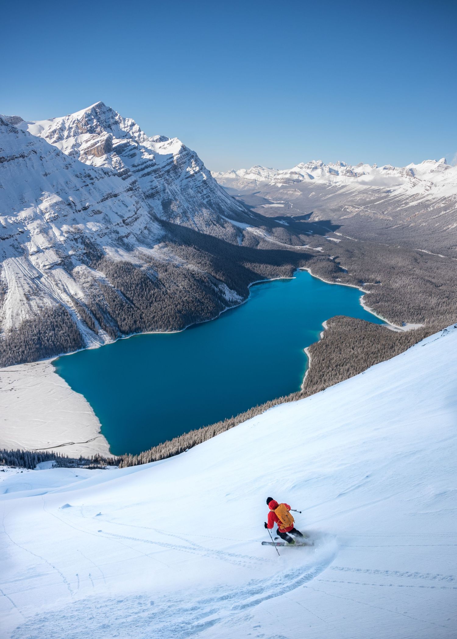 Skier descends snowy slope above turquoise mountain lake surrounded by snow-capped peaks.
