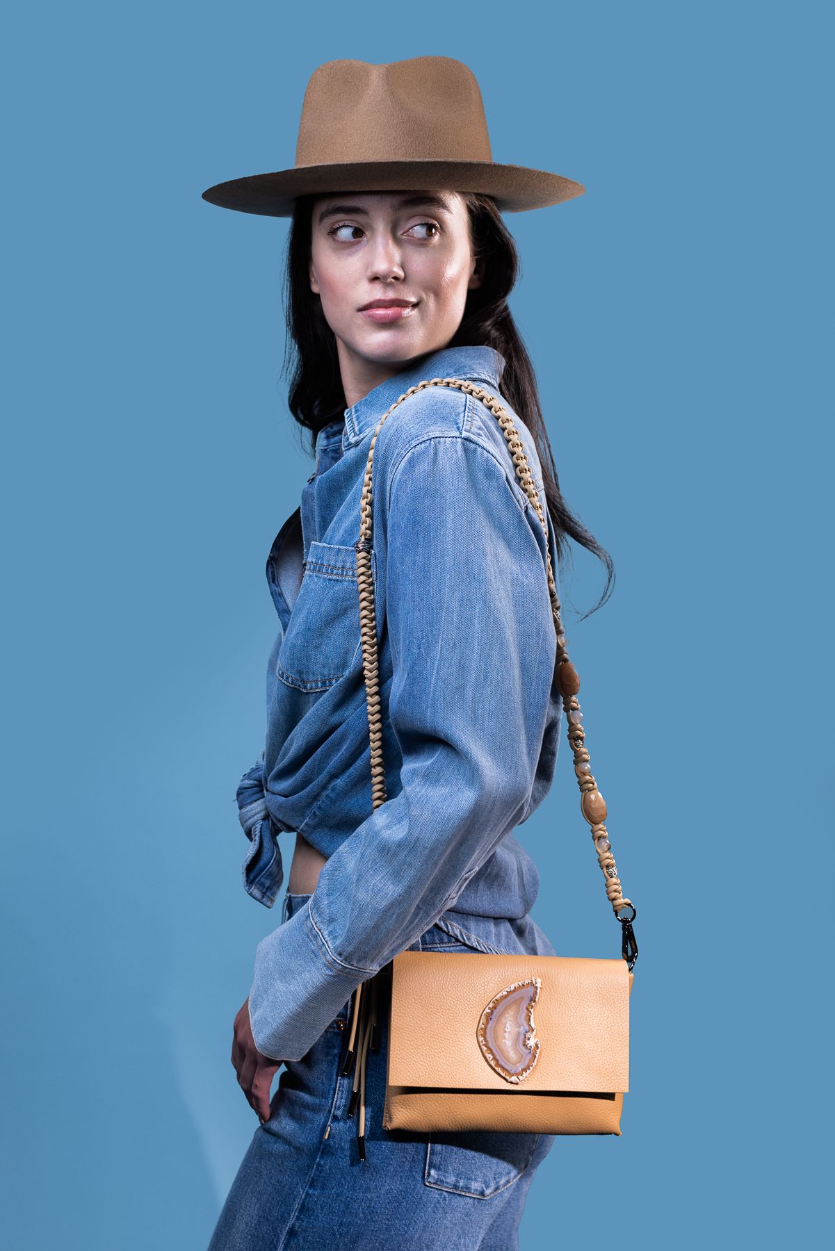Model wearing denim jacket and brown hat with tan leather crossbody bag poses against blue background.