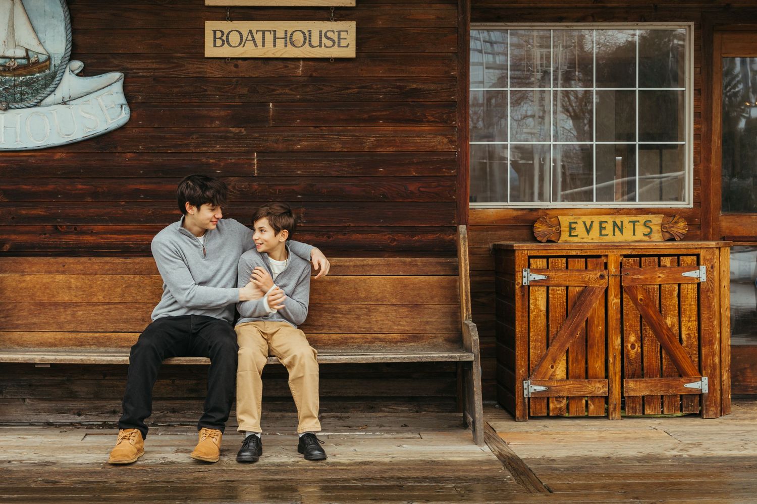 Two brothers sit close together on a wooden bench outside a rustic boathouse with event rental signage.