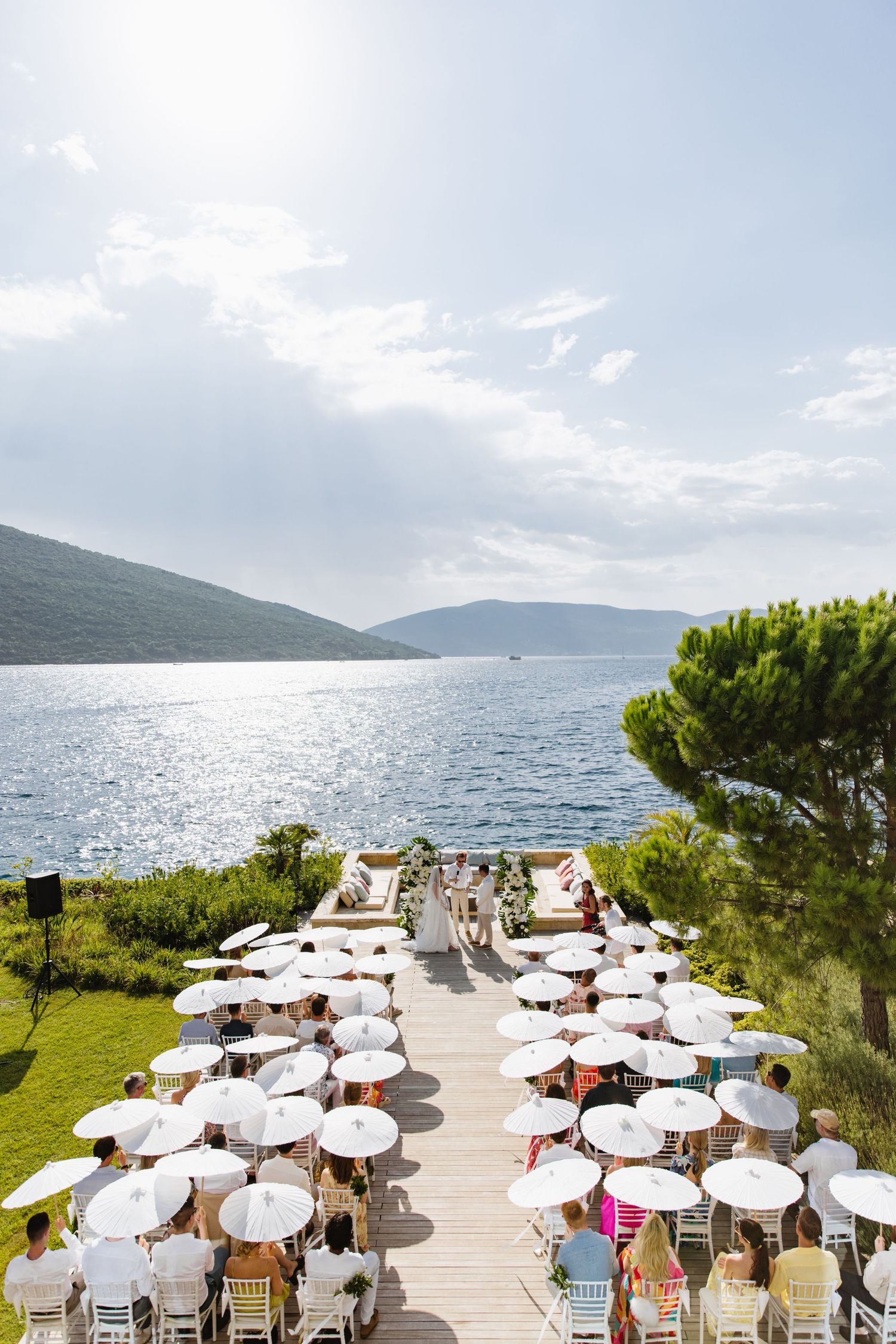 Outdoor wedding ceremony setup with white chairs and umbrellas overlooking a scenic mountain lake on a sunny day.