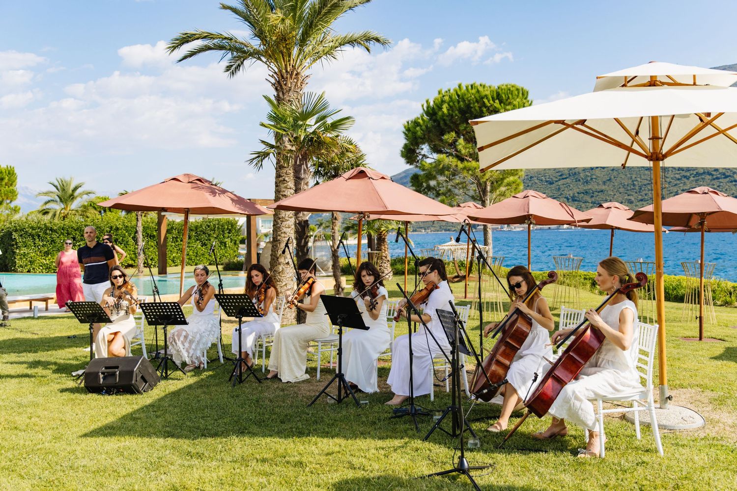 Musicians perform at an outdoor wedding ceremony with umbrellas and palm trees by the ocean on a sunny day.