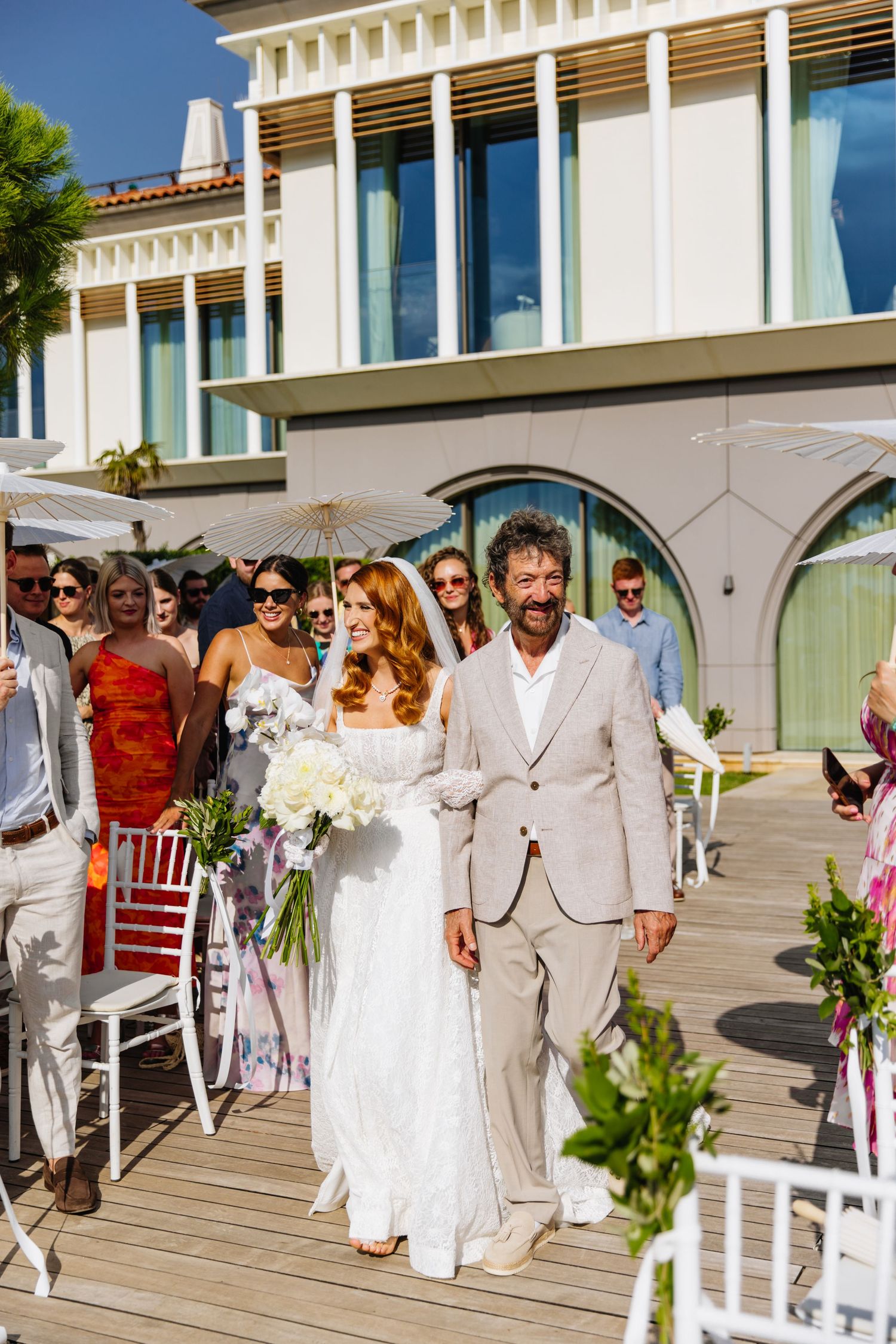 A couple walks down a wooden deck during a beachside wedding ceremony with guests seated on white chairs.
