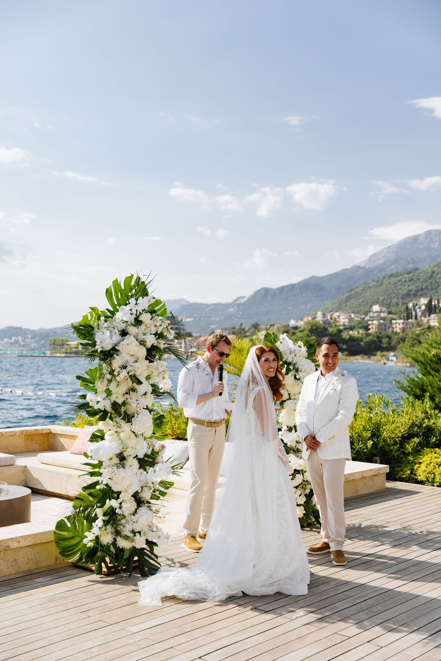 Beautiful outdoor wedding ceremony overlooking a lake and mountains, decorated with white floral arch and monstera leaves.