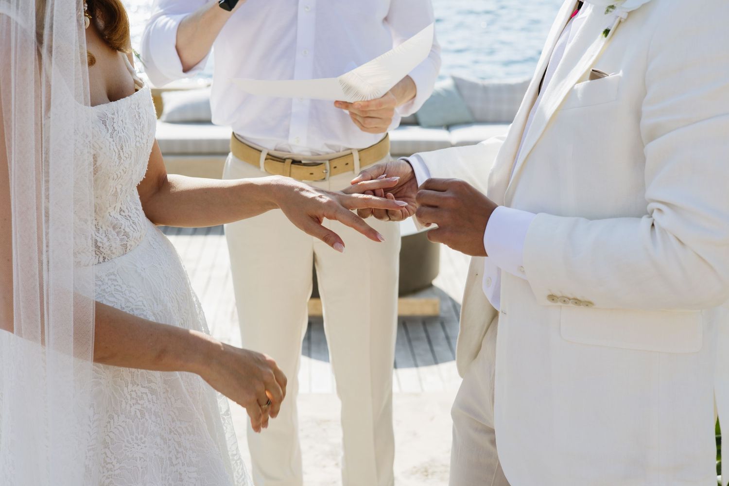 Close-up shot of hands exchanging wedding rings during an outdoor ceremony in white attire.