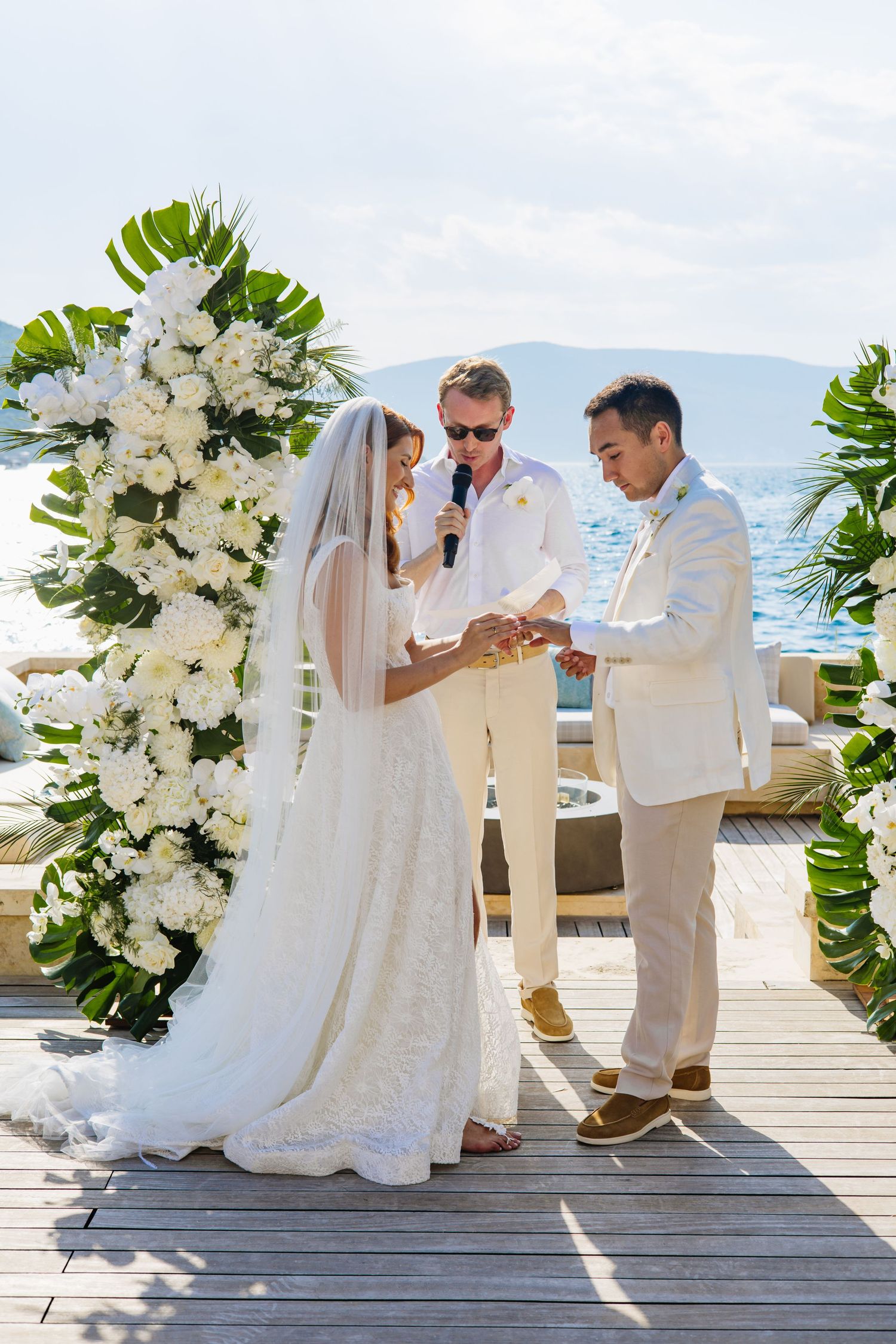 Elegant wedding ceremony on a deck with tropical backdrop and white floral arch decorations on a sunny day.