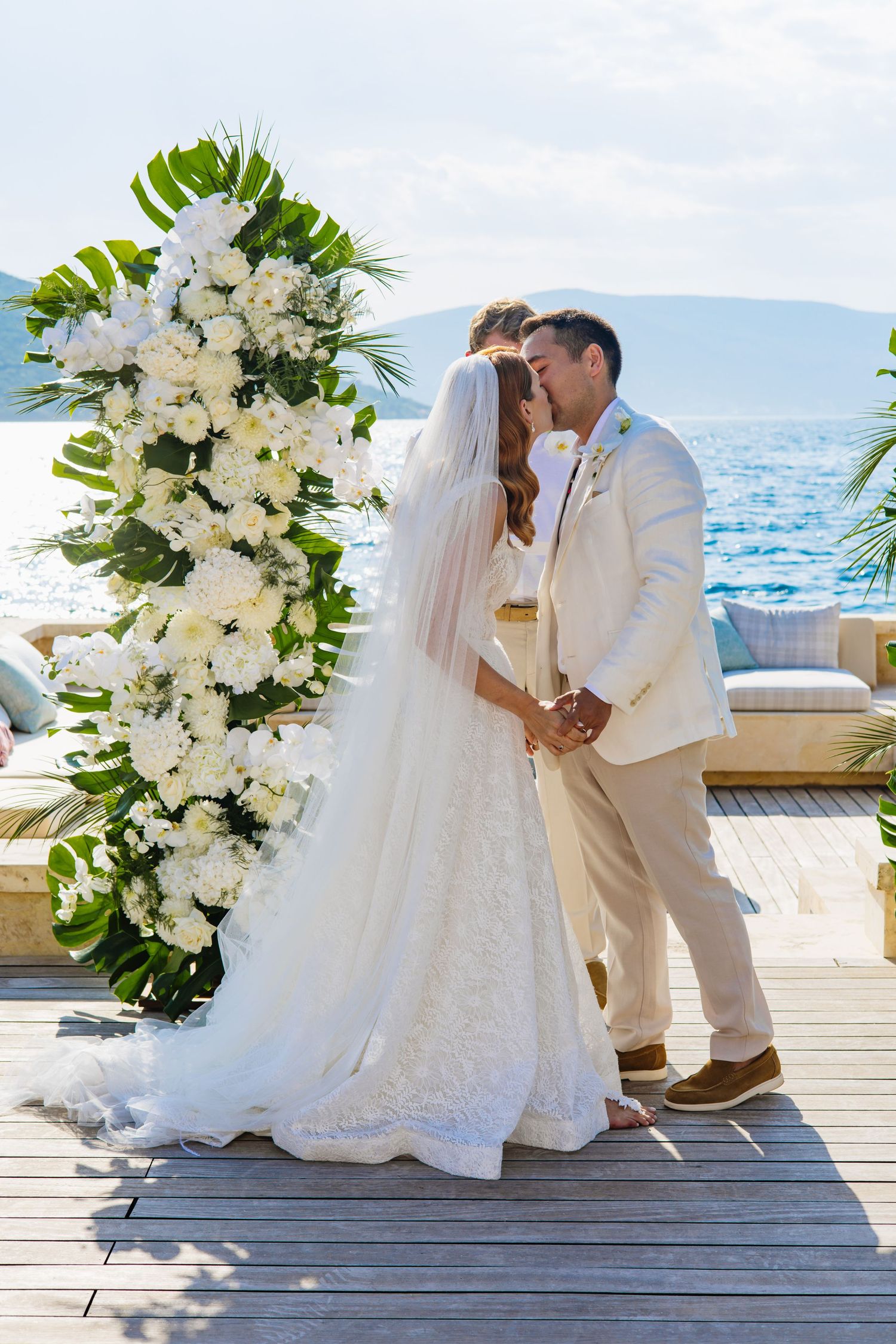 A beachside wedding ceremony setup with white floral arrangements and tropical leaves on a wooden deck overlooking the ocean.