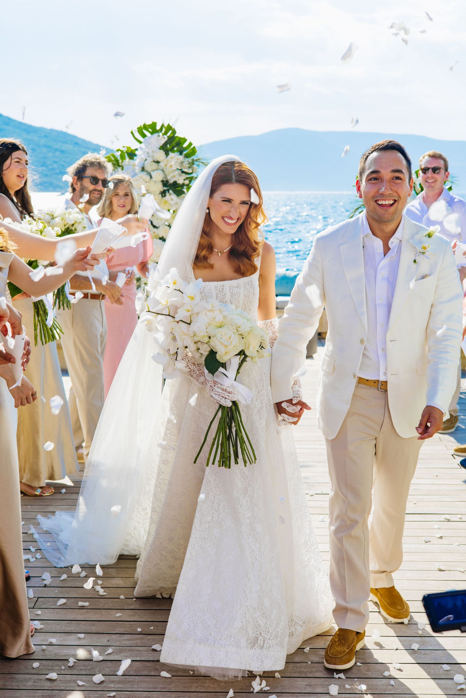 Wedding party celebrates on scenic lakeside dock with flower petals and decorative arch during summer ceremony.
