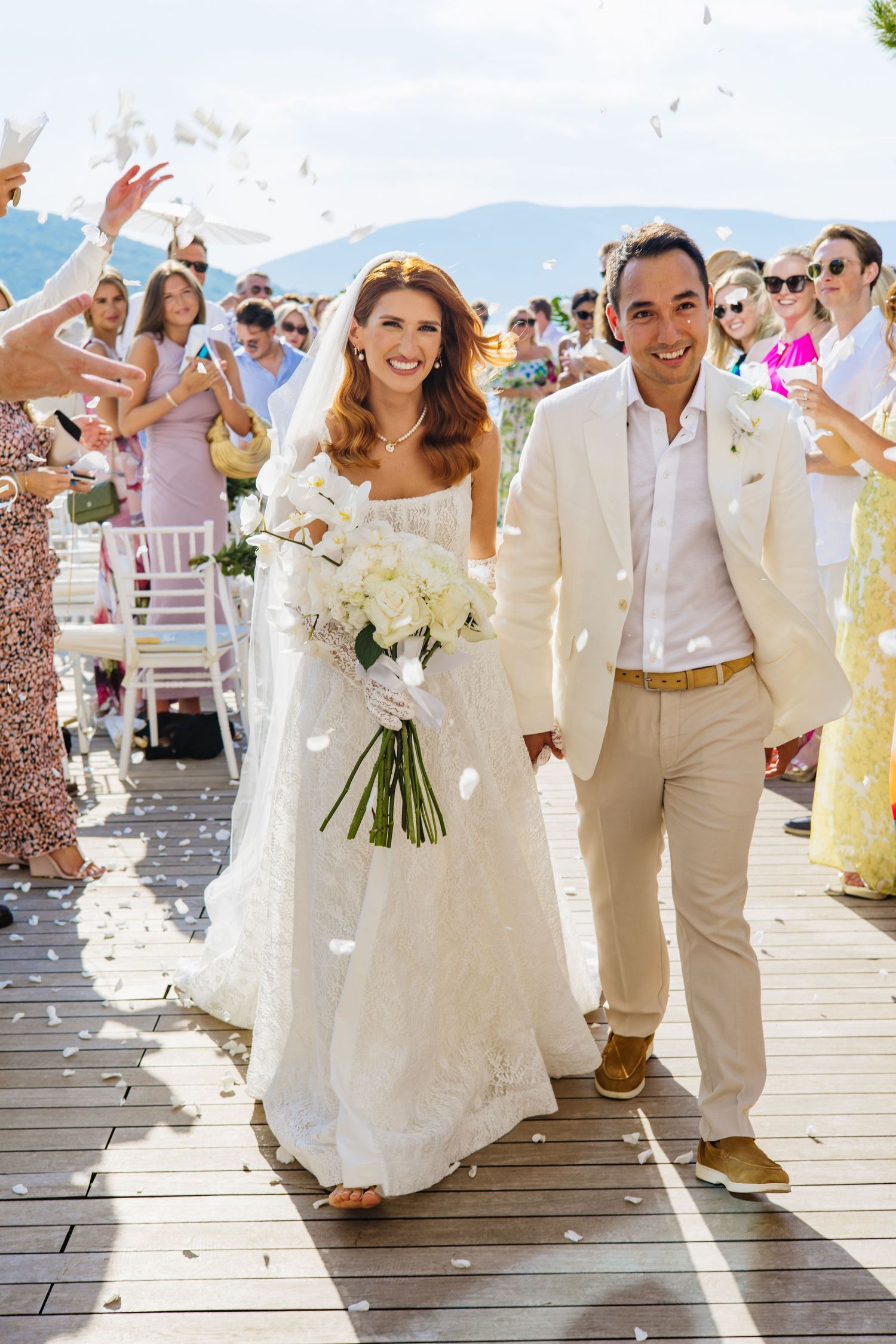 Happy couple walks down aisle during outdoor wedding ceremony as guests throw flower petals in celebration.
