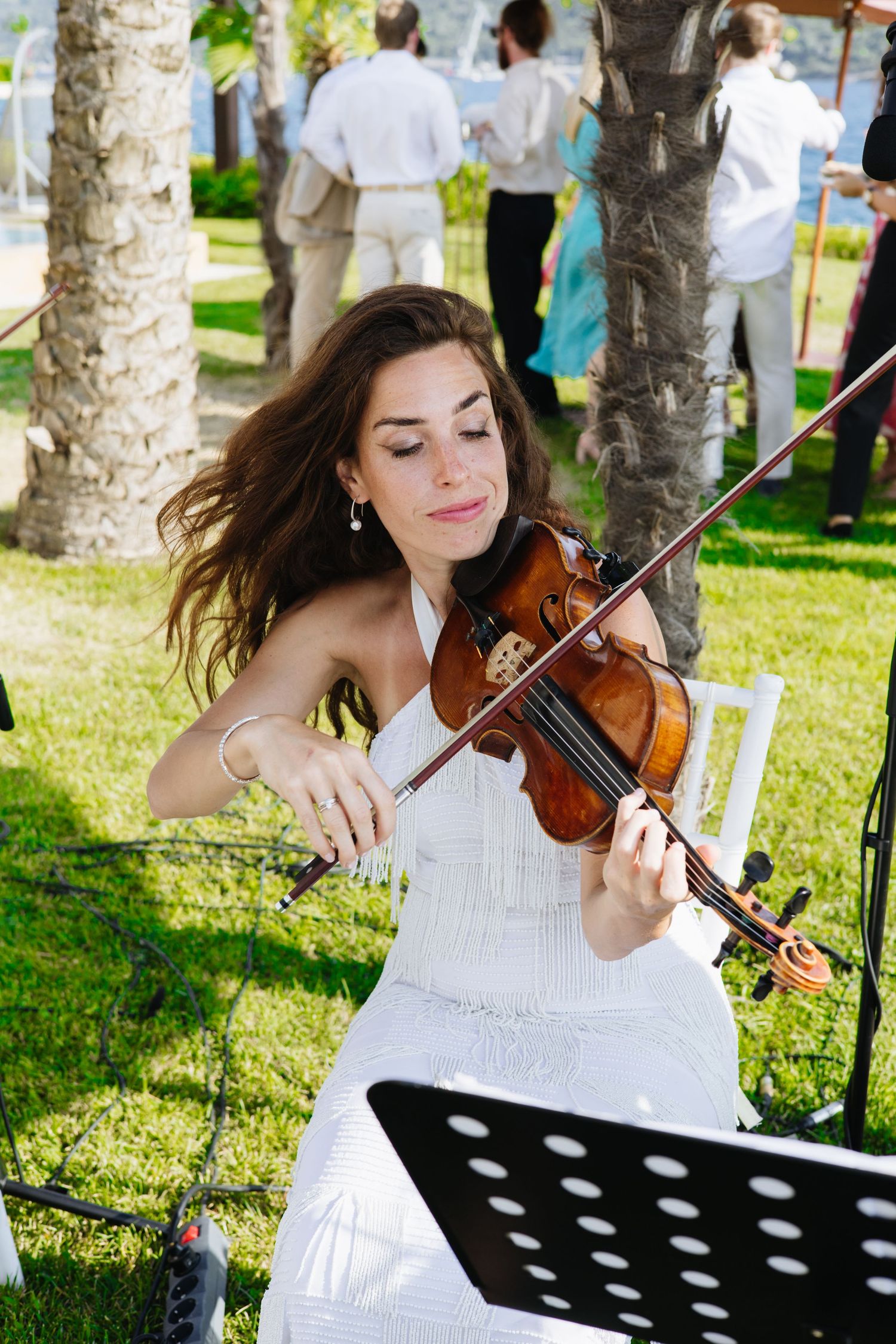 A musician in a white dress plays violin at an outdoor event with trees in the background.