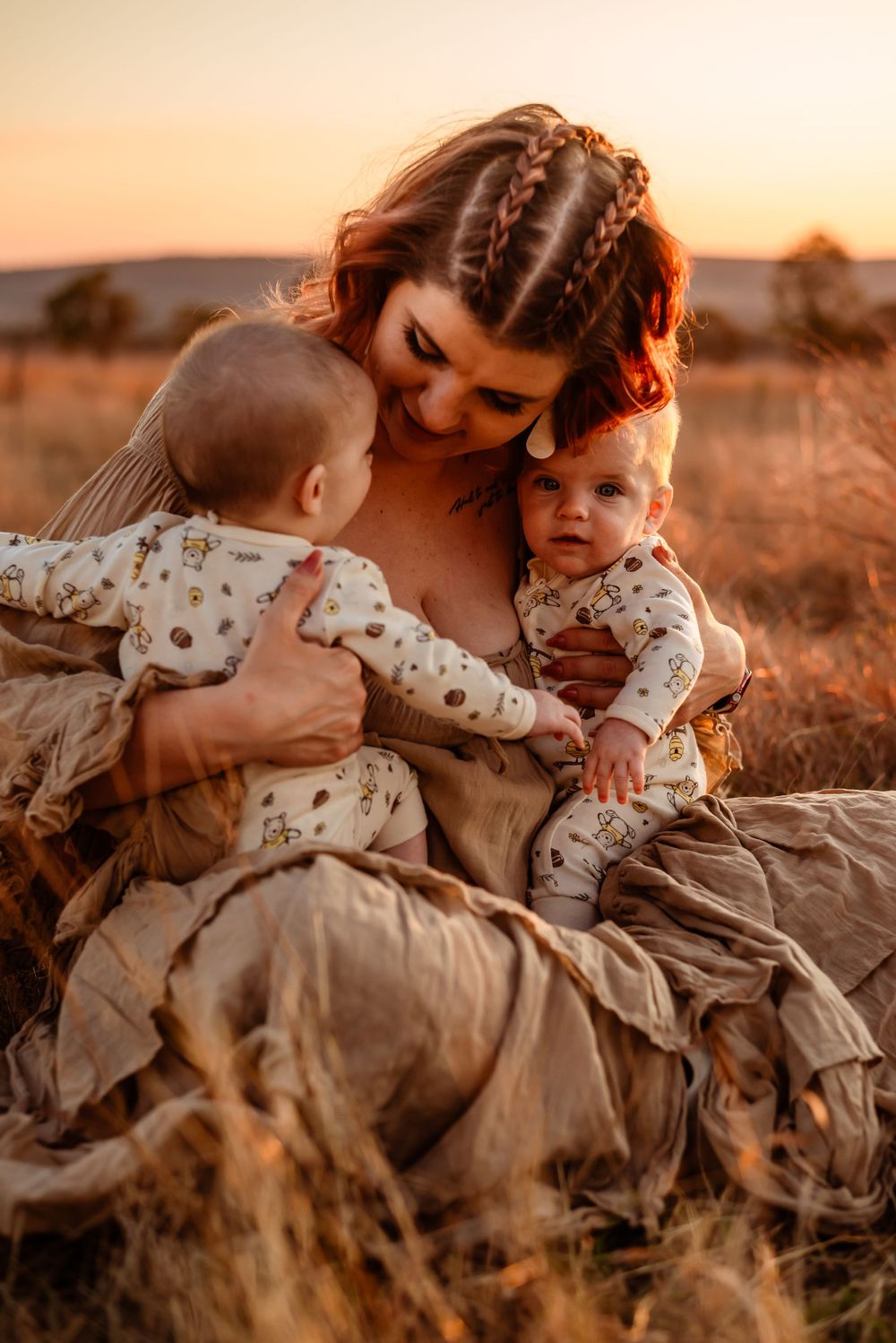 Mother embracing twin babies in matching outfits during golden hour sunset.