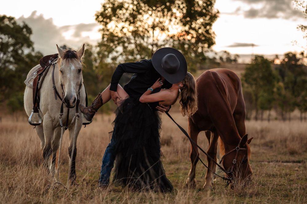 Two horses standing in dry grassland at sunset with someone in a cowboy hat between them.